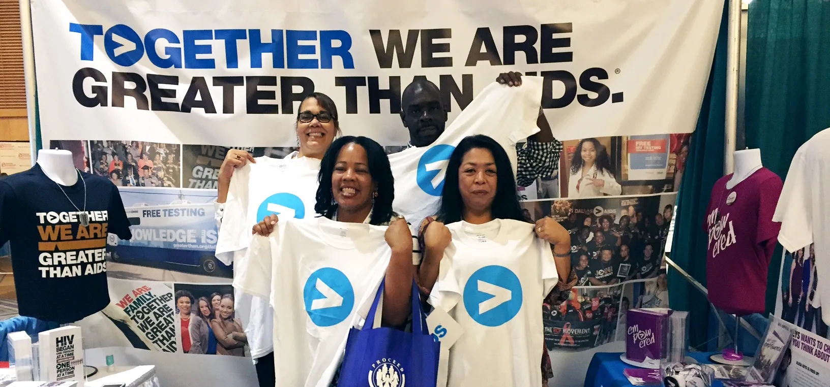 Four people smiling at Greater Than HIV at USCA 2015 holding t-shirts with "Greater Than" symbol